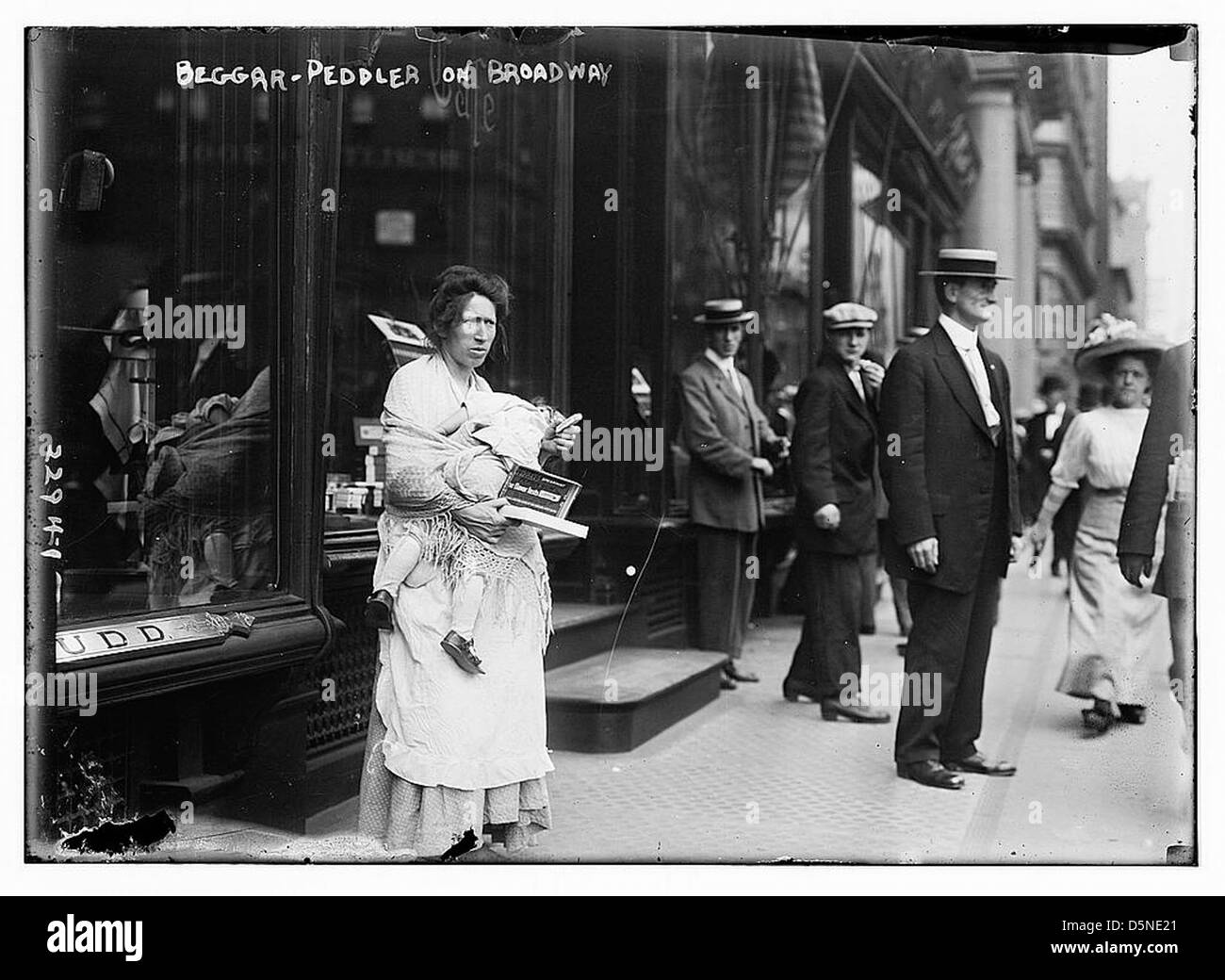 Una fotografia degli anni '1910 che raffigura un venditore di mendicanti a Broadway a New York. L'immagine mostra una donna che tiene in braccio un bambino, con un uomo che indossa un cappello, possibilmente una pannellatrice, nell'ambiente urbano frenetico. La scena evidenzia la povertà e la lotta degli individui a New York durante questo periodo di tempo. Foto Stock