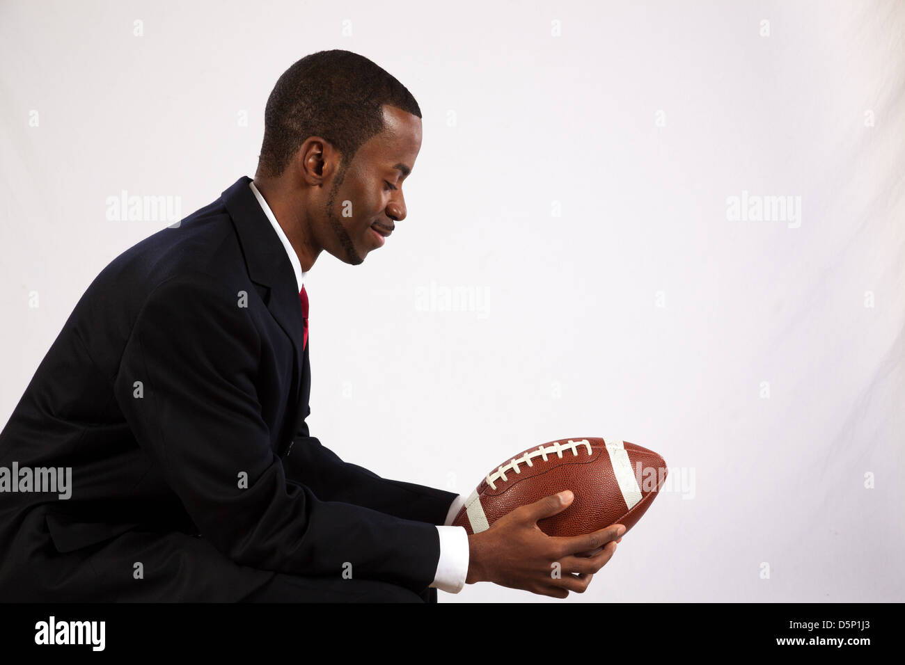 Bel uomo nero che indossa un abito scuro, una camicia bianca e cravatta rossa, tenendo un football americano e ricordando la sua gioventù Foto Stock