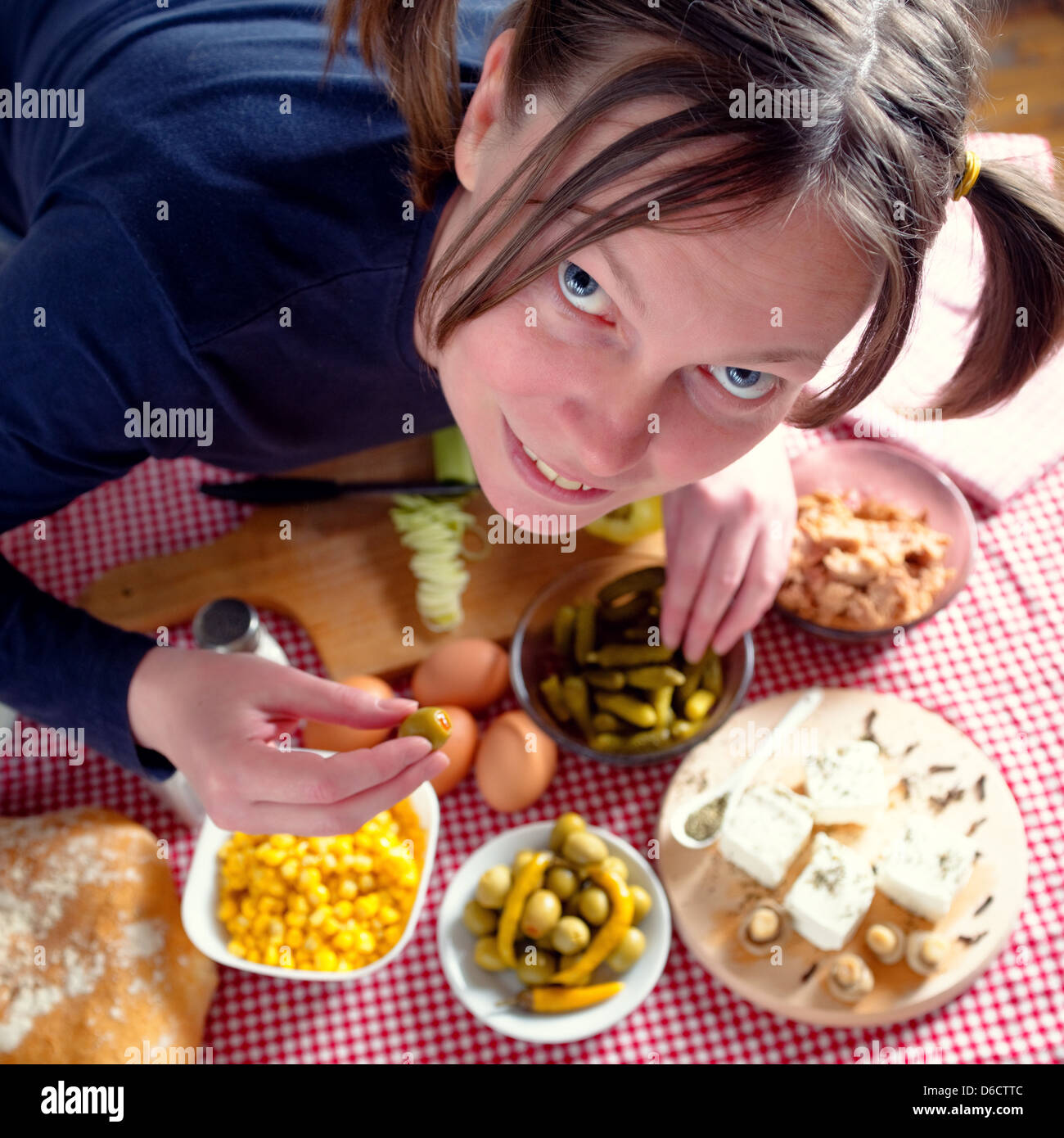 Donna di mangiare olive. Bella Donna sorridente con pig-tail mangiare olive. Tavolo da cucina con freschi ingredienti alimentari. Foto Stock