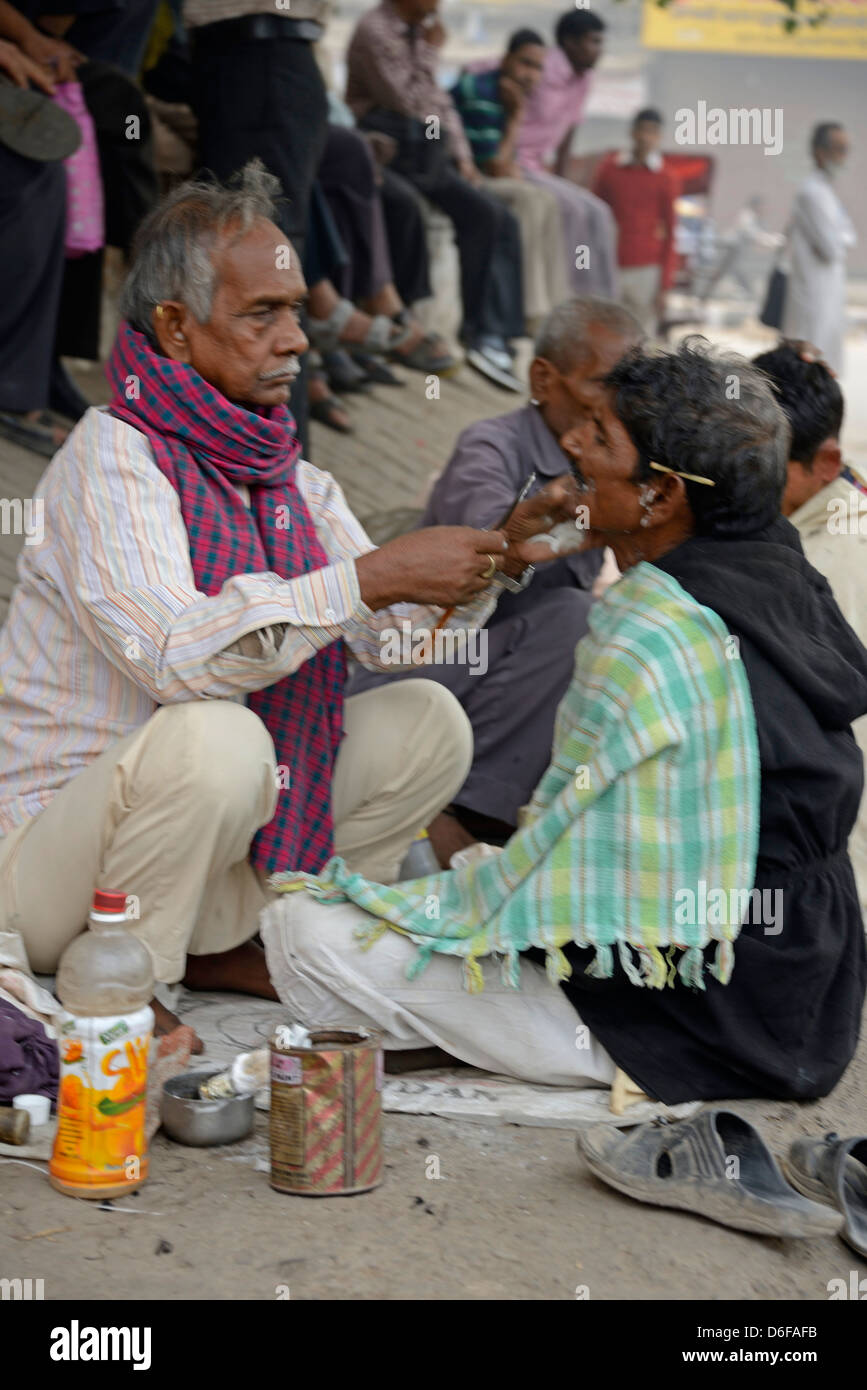 Un barbiere che dà al suo cliente una rasatura bagnata tra una fila di barbieri che squattano in una strada nella vecchia Delhi, India. Foto Stock