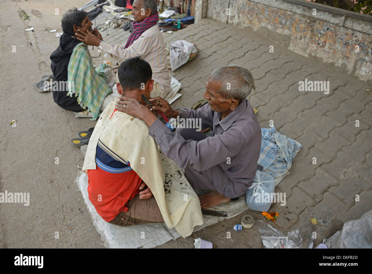 Un barbiere che dà al suo cliente una rasatura bagnata tra una fila di barbieri che squattano in una strada nella vecchia Delhi, India. Foto Stock