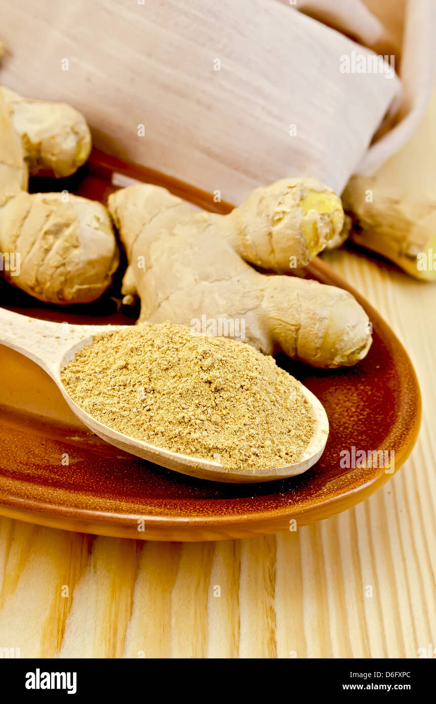 Radici di zenzero fresco, un cucchiaio di legno con una polvere di zenzero su ceramica, stoffa contro un pannello di legno Foto Stock