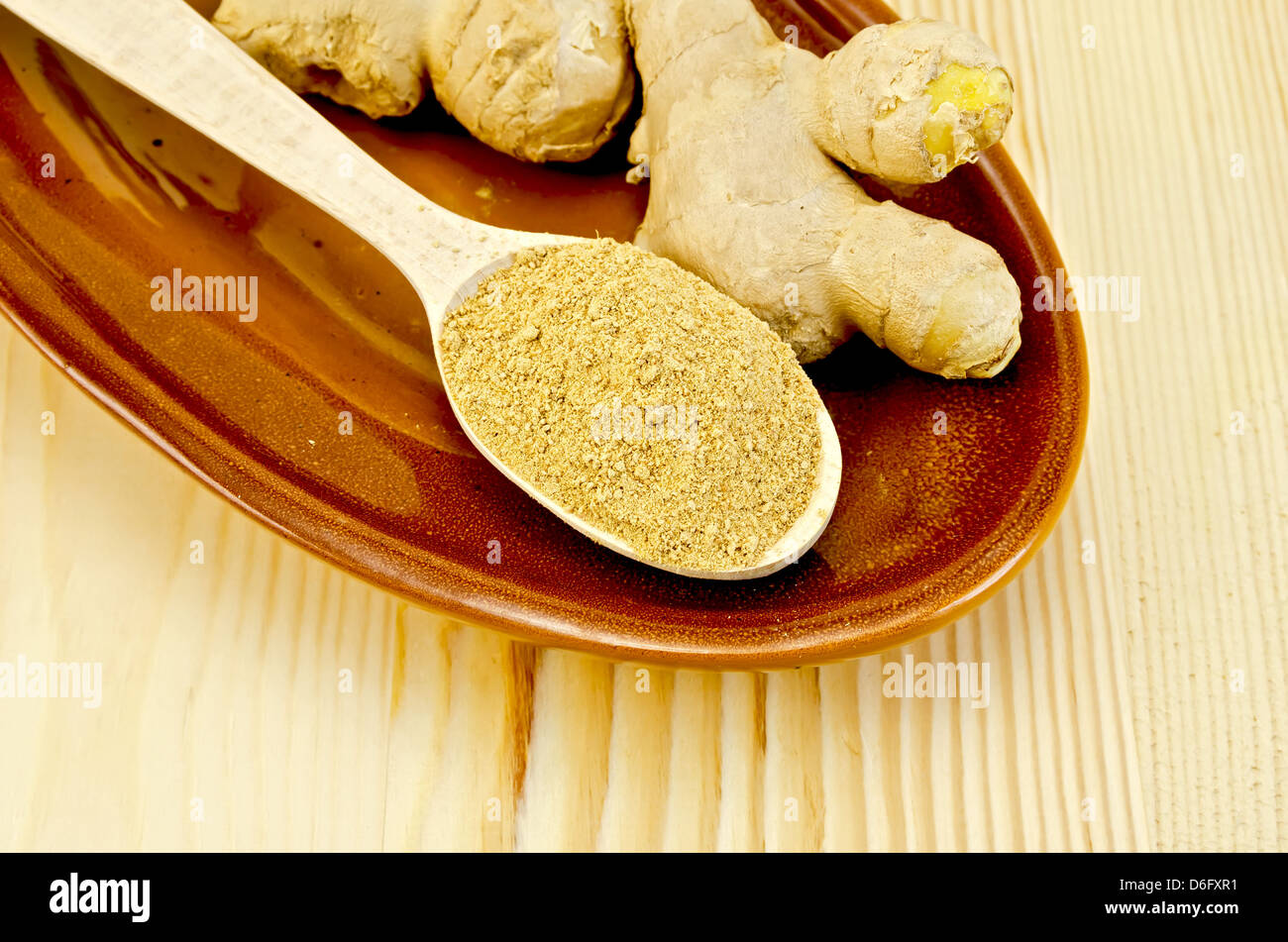 Radici di zenzero fresco, un cucchiaio di legno con una polvere di zenzero su vasellame contro un pannello di legno Foto Stock