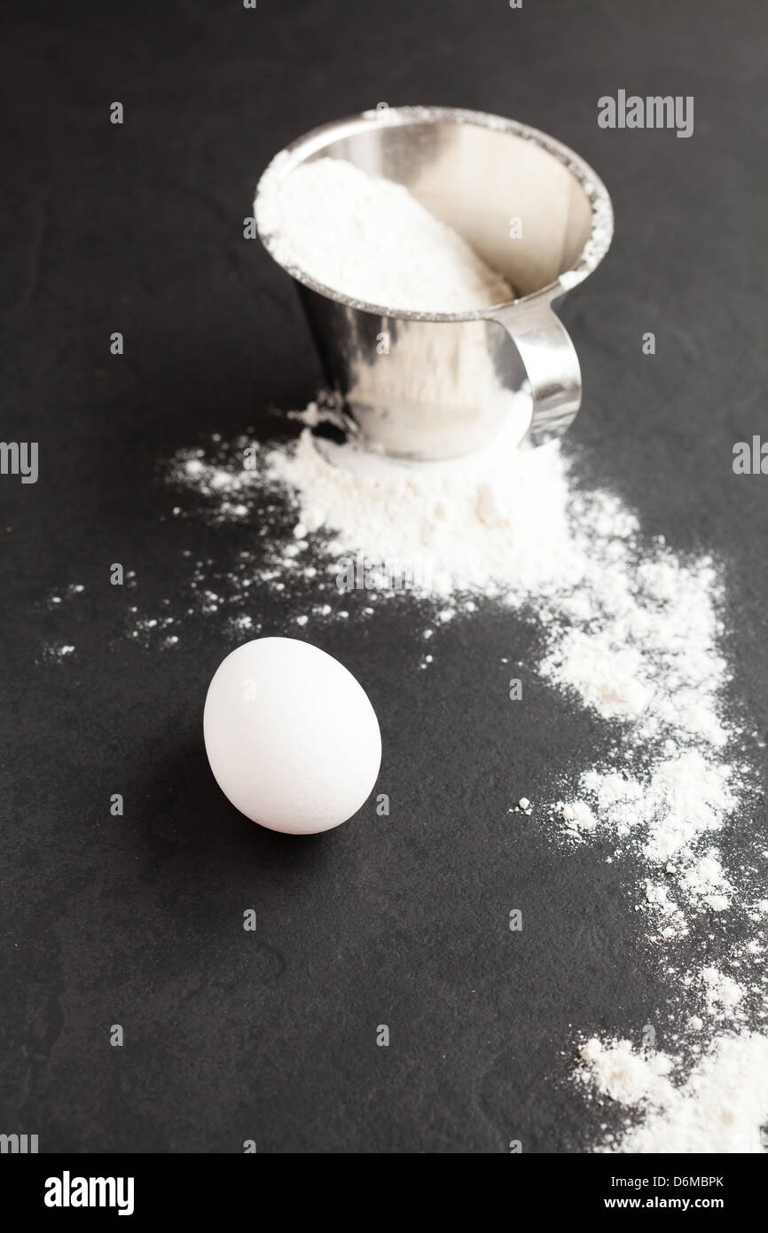 Uovo di pollo con la farina in metallo coppa di misurazione e diffuso su sfondo scuro Foto Stock