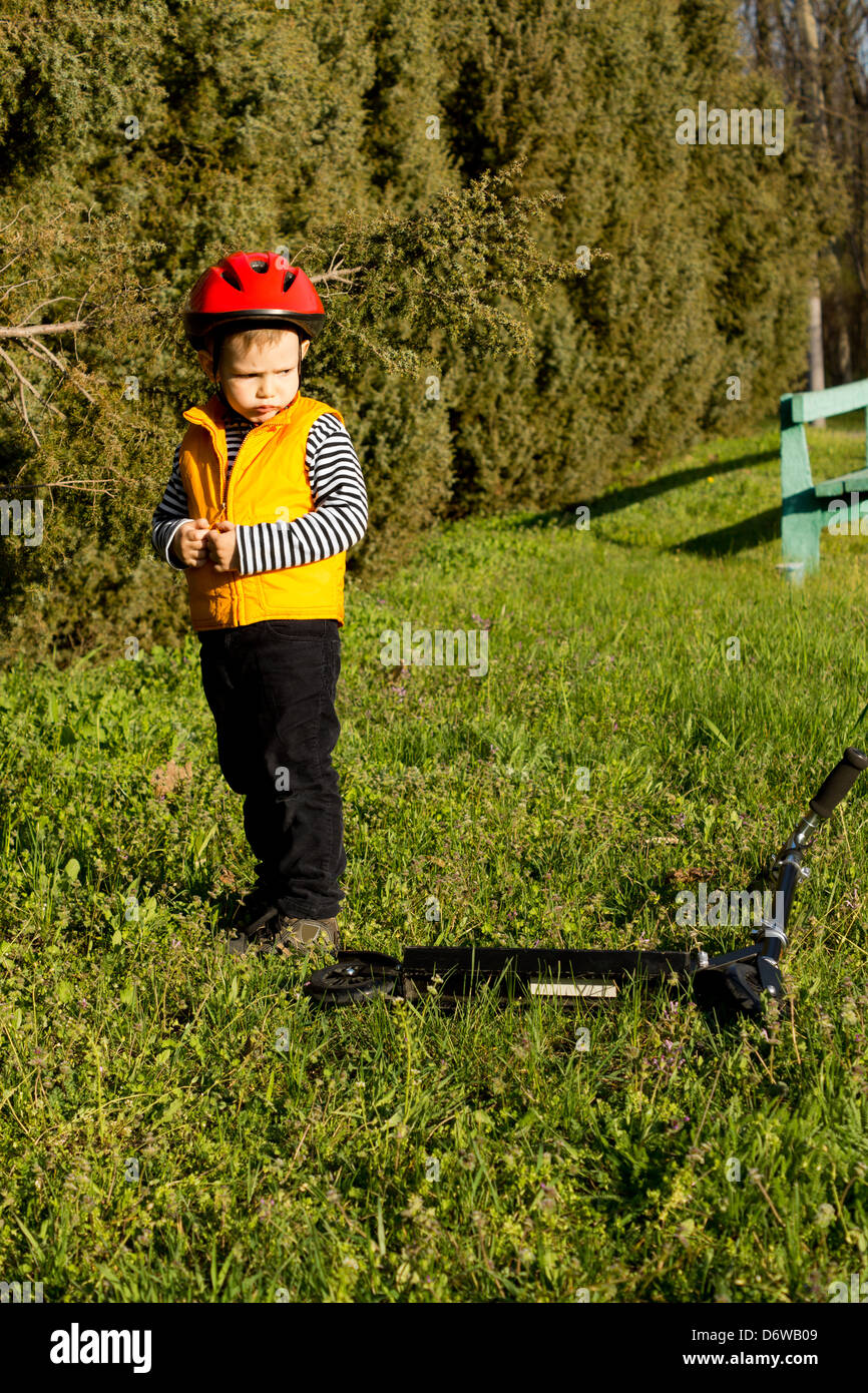 Sulky ragazzino con un broncio permanente di espressione guardando sopra la sua spalla al suo scooter giacente in erba verde nella luce del sole serale. Foto Stock