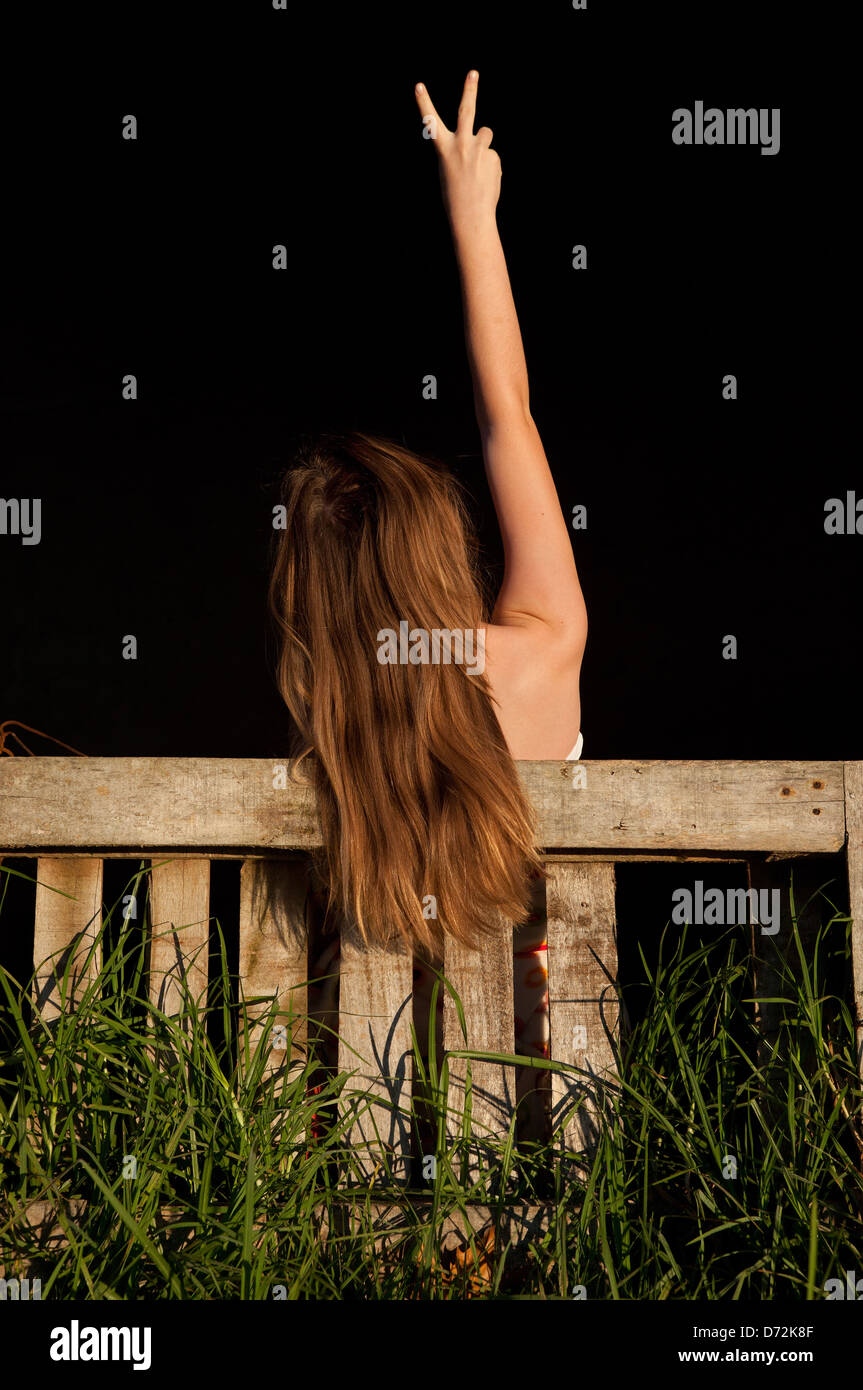 Ragazza giovane con il suo braccio in aria dando un segno di vittoria con capelli lunghi appesa sopra una fattoria cancello di legno ed erba selvatica crescente Foto Stock