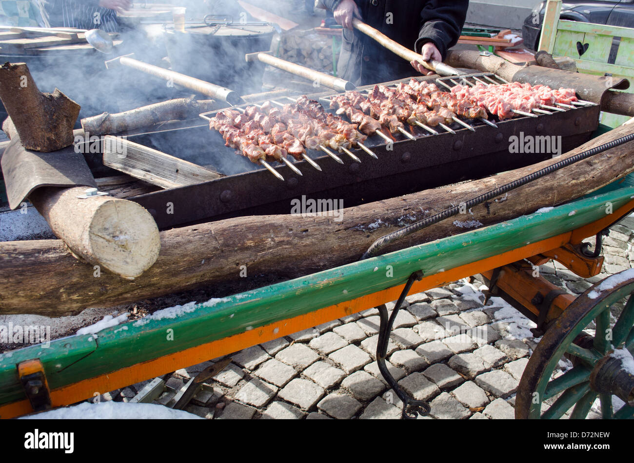 Man mano con forcella cuocere la carne di maiale sul fuoco in outdoor street evento. preparazione alimentare industria. la gente a piedi di distanza. fumo Foto Stock