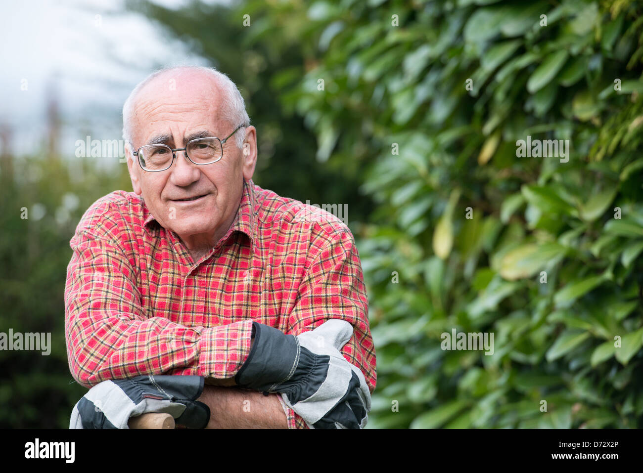 Uomo anziano in piedi in giardino, indossare abiti da lavoro e azienda vanga Foto Stock