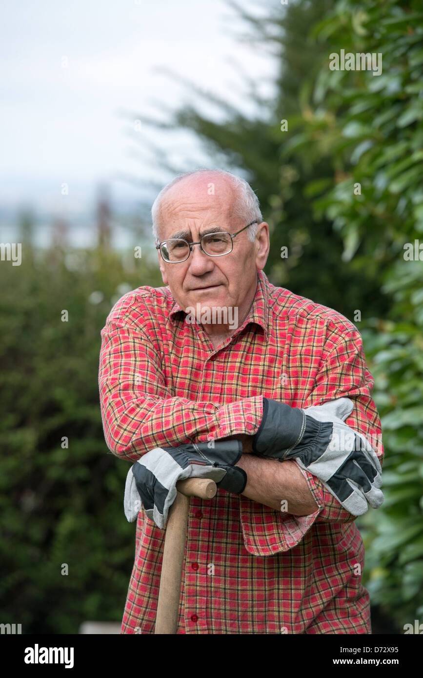 Uomo anziano in piedi in giardino, indossare abiti da lavoro e azienda vanga Foto Stock