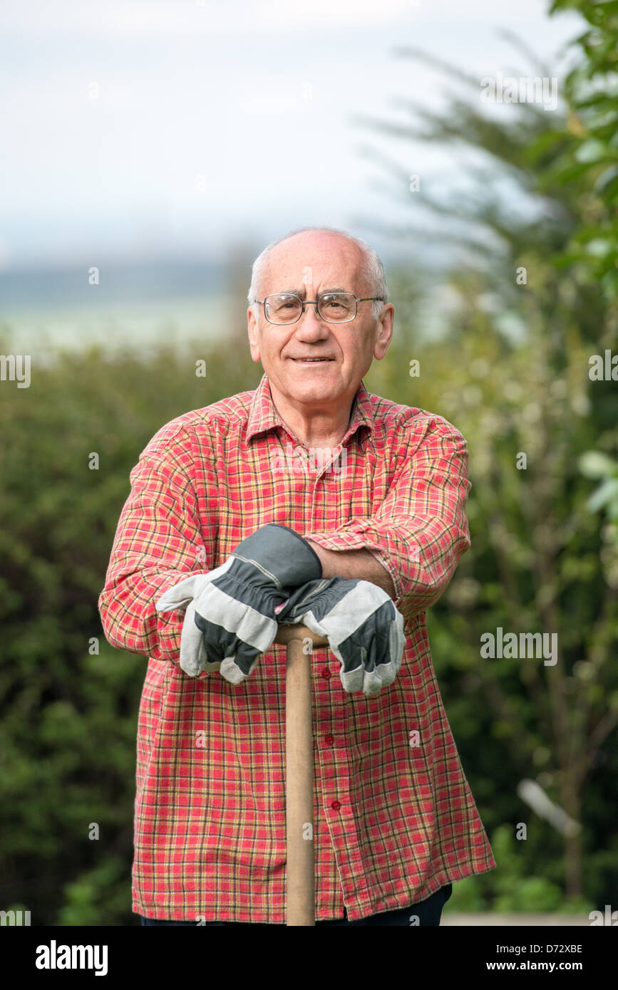 Uomo anziano in piedi in giardino, indossare abiti da lavoro e azienda vanga Foto Stock