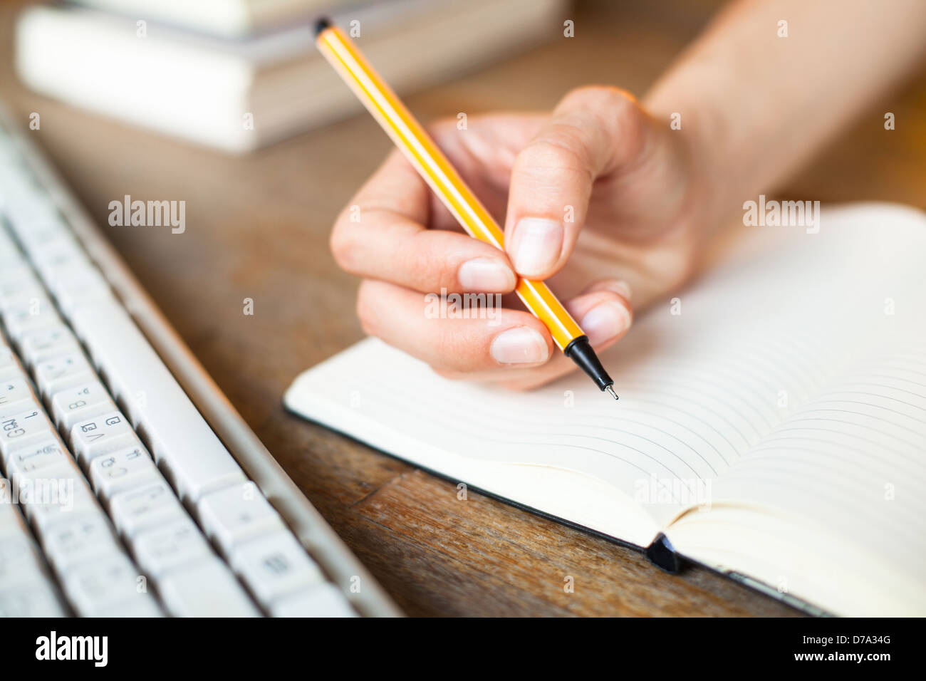 Mani scrive una penna in un notebook, la tastiera del computer e una pila di libri in background. Foto Stock