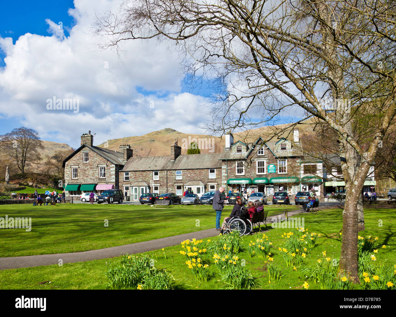 La gente seduta sulle panchine intorno al villaggio verde godendo il sole primaverile in Grasmere Cumbria Inghilterra UK GB EU Europe Foto Stock