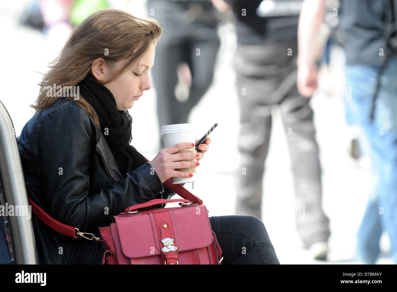 Una ragazza si siede su un banco con un caffè in mano mentre si utilizza un telefono. Foto Stock