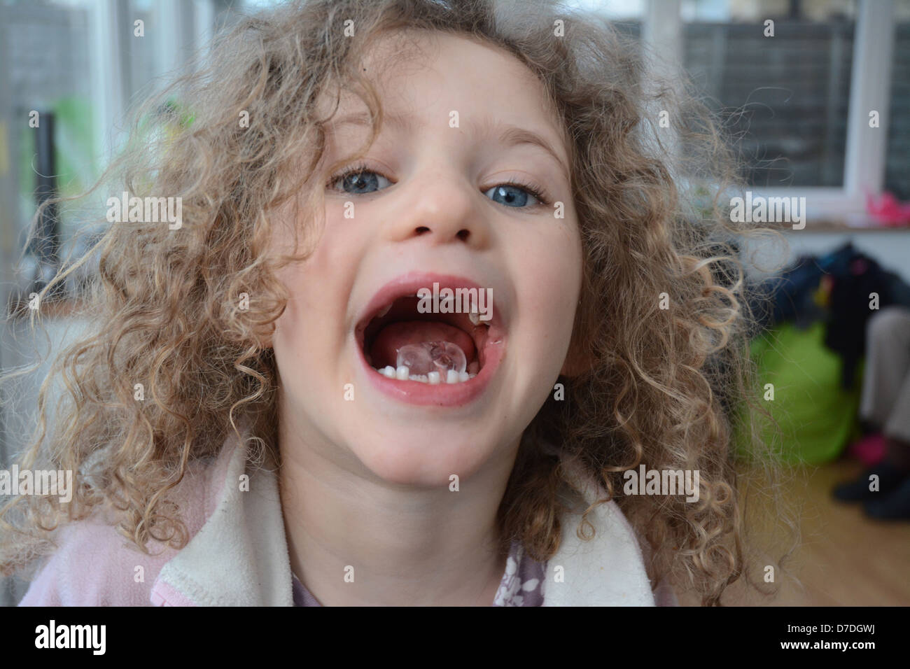 Bambina di cinque anni con i capelli ricci e gli occhi blu che mostra un cubetto di ghiaccio nella sua bocca aperta Foto Stock