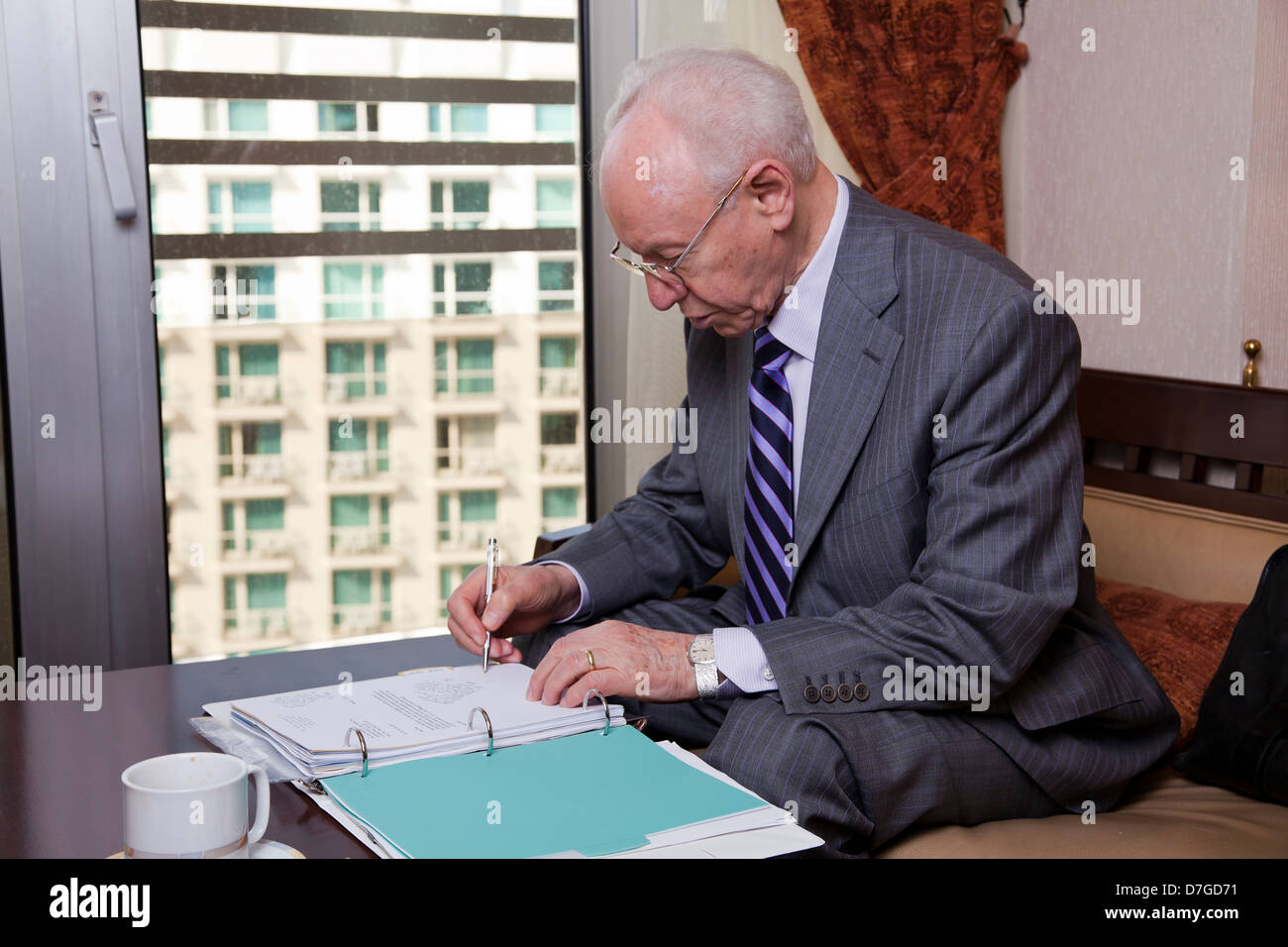 Un anziano (nella sua 80's) business man tuta indossa cravatta seduti in hotel della business lounge andando su alcune carte scrivere note Foto Stock