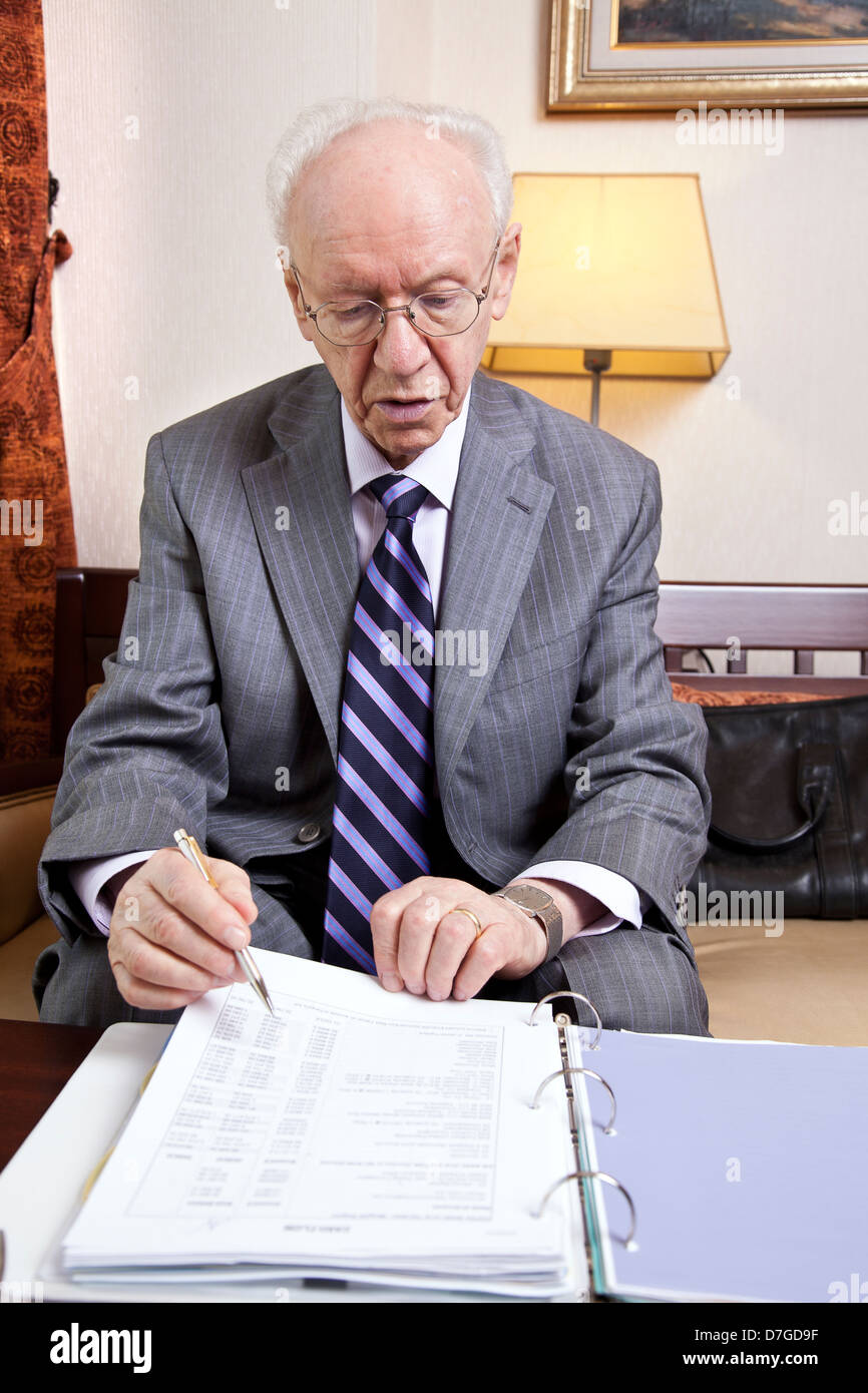 Un anziano (nella sua 80's) business man tuta indossa cravatta seduti in hotel della business lounge andando su alcune carte i dettagli di bilancio. Foto Stock