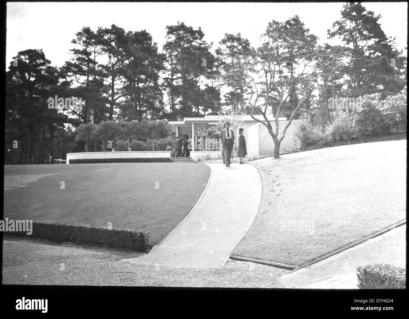Questa immagine mostra un gruppo di persone di fronte a una casa "moderna", con un percorso in mattoni che taglia il prato, riflettendo il design architettonico suburbano della metà del XX secolo e le riunioni sociali intorno a nuovi stili abitativi. Foto Stock