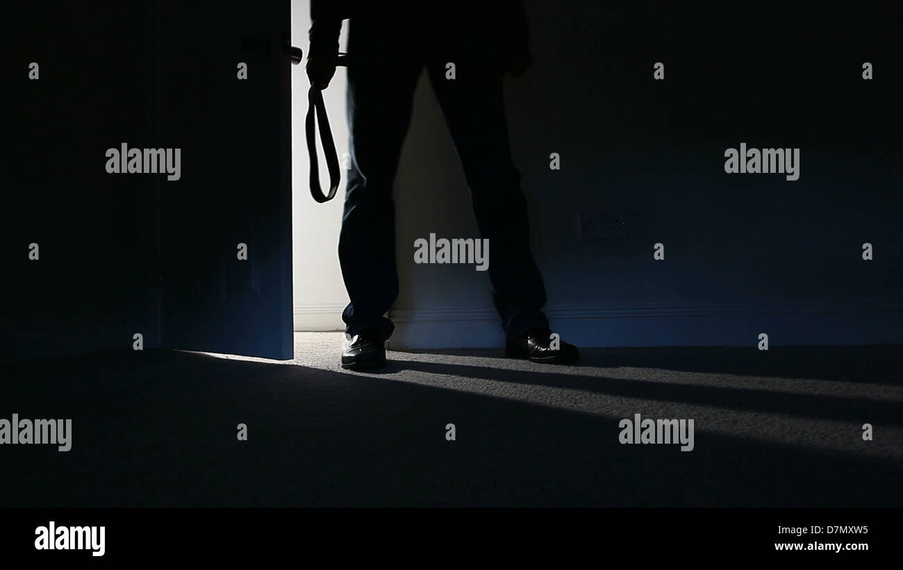 Silhouette di un uomo in piedi in una stanza buia, tenendo una cinghia attorno a battere qualcuno con esso, lo streaming di luce attraverso una porta aperta. Foto Stock