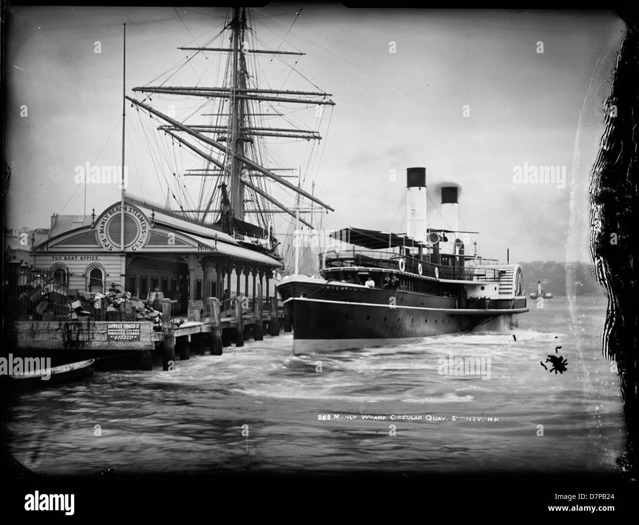 Un'immagine storica in bianco e nero del Manly Wharf e del Circular Quay a Sydney, Australia, con il Powerhouse Museum visibile sullo sfondo. La scena include un grande albero di legno proveniente da una nave, nonché un piroscafo a pale attraccato al molo. L'immagine cattura il vivace porto con le navi, tra cui il traghetto di Manly, attraccato al molo. Foto Stock