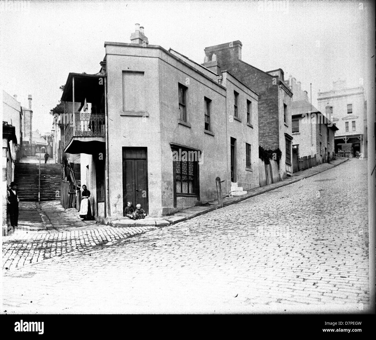 Questa fotografia in bianco e nero raffigura una storica scena di strada a The Rocks, Sydney, che mostra la vivace vita quotidiana in una zona acciottolata. L'immagine include punti di riferimento chiave come Hit o Miss Hotel, che forniscono uno sguardo sulla storia culturale e architettonica della zona. Foto Stock