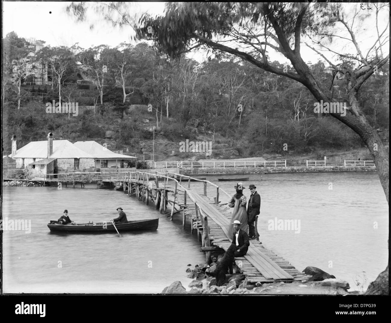 Una fotografia storica di un ponte di legno sulla Mosman Bay, Australia, cattura una tranquilla scena lungo il fiume con un gruppo di persone e un cane sulla riva. L'area era nota per le sue bellezze naturali e l'attività marittima. Foto Stock