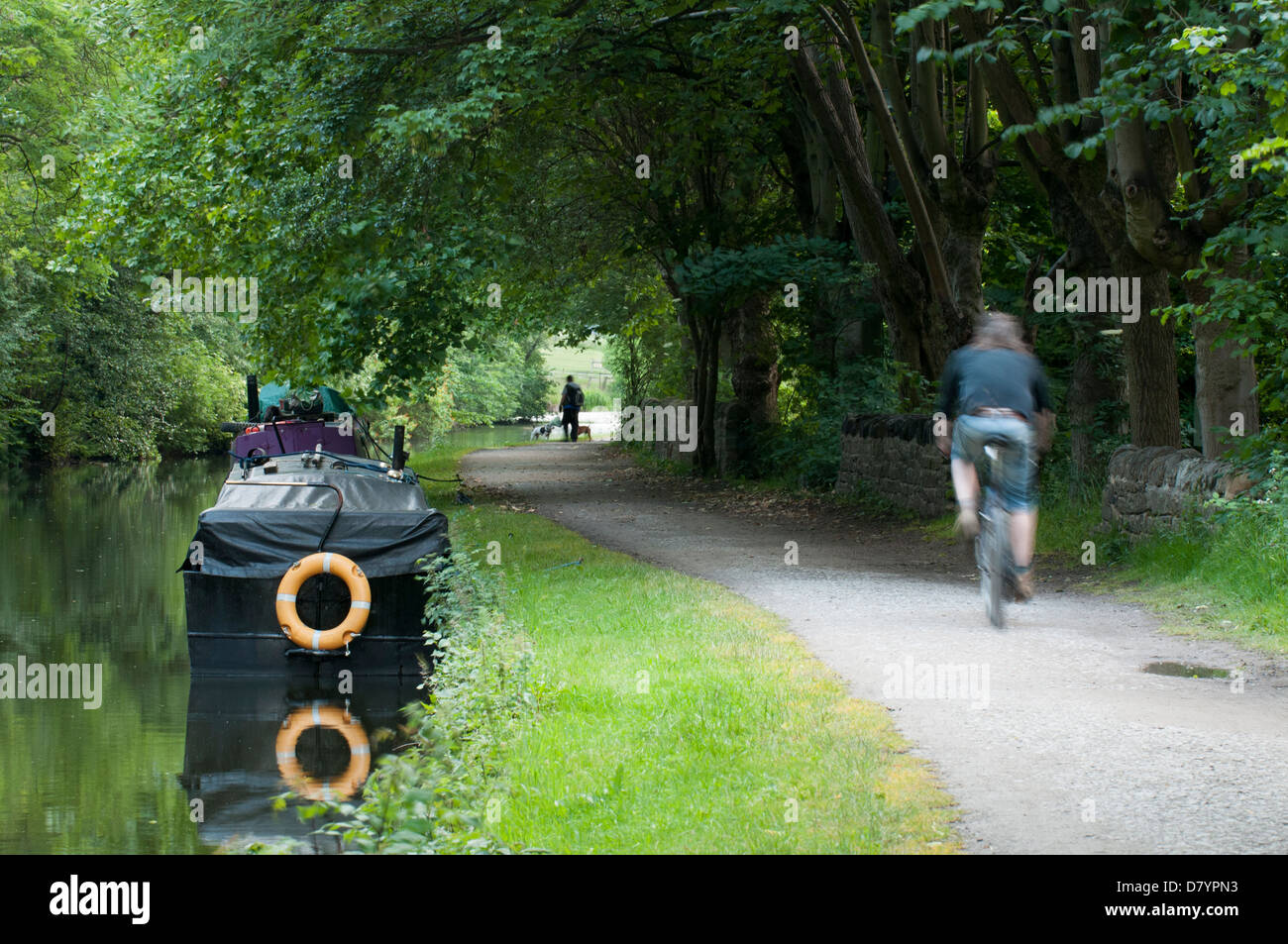 Silenzioso, Scenic, rurale, viale alberato di tratto di strada alzaia con barche ormeggiate & Man in bicicletta lungo - Leeds Liverpool Canal, Saltaire, nello Yorkshire, Inghilterra, Regno Unito. Foto Stock