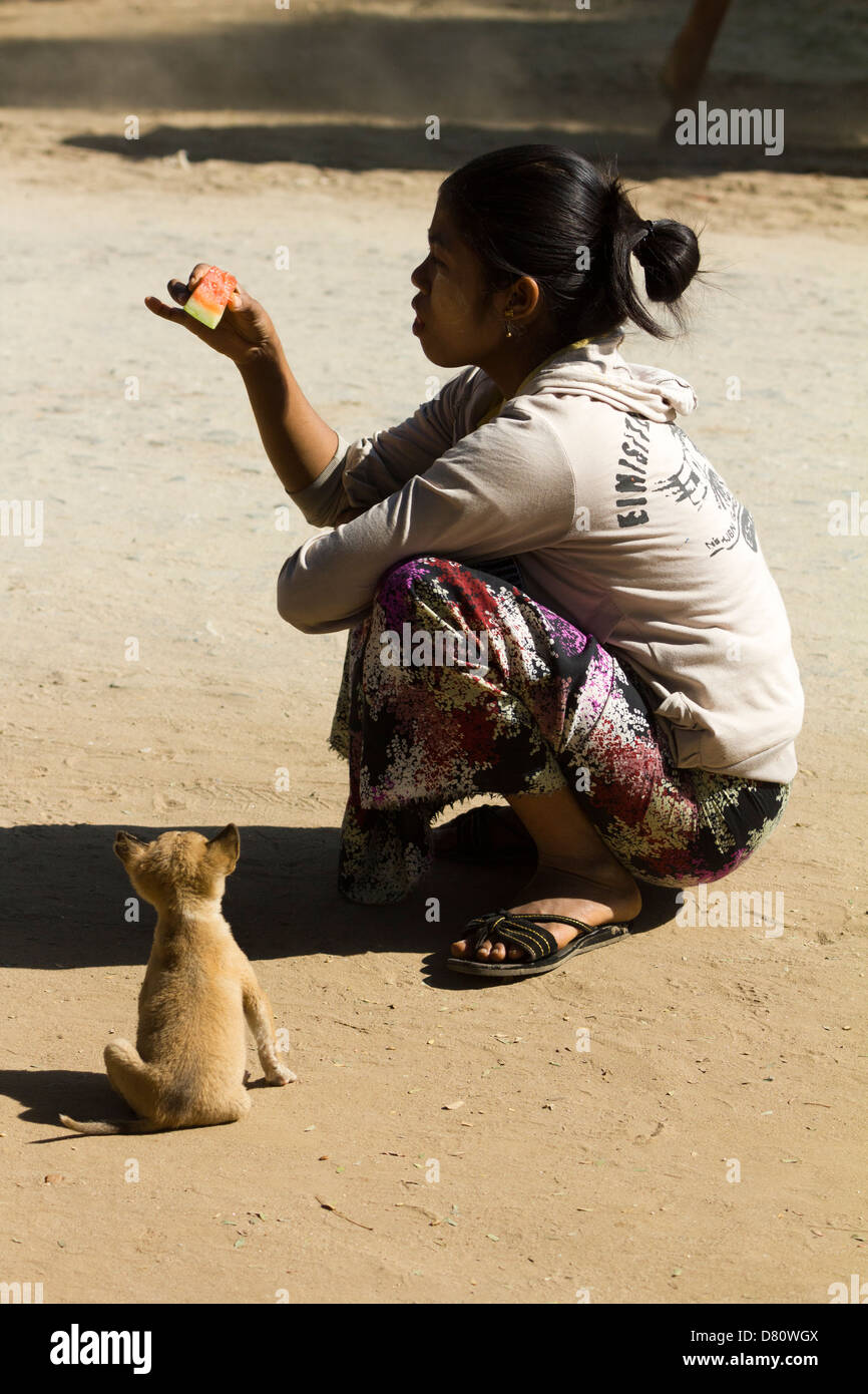 Una donna mangiare anguria in mezzo alla strada mentre un minuscolo cucciolo guarda con speranza - Mingun, Myanmar 3 Foto Stock