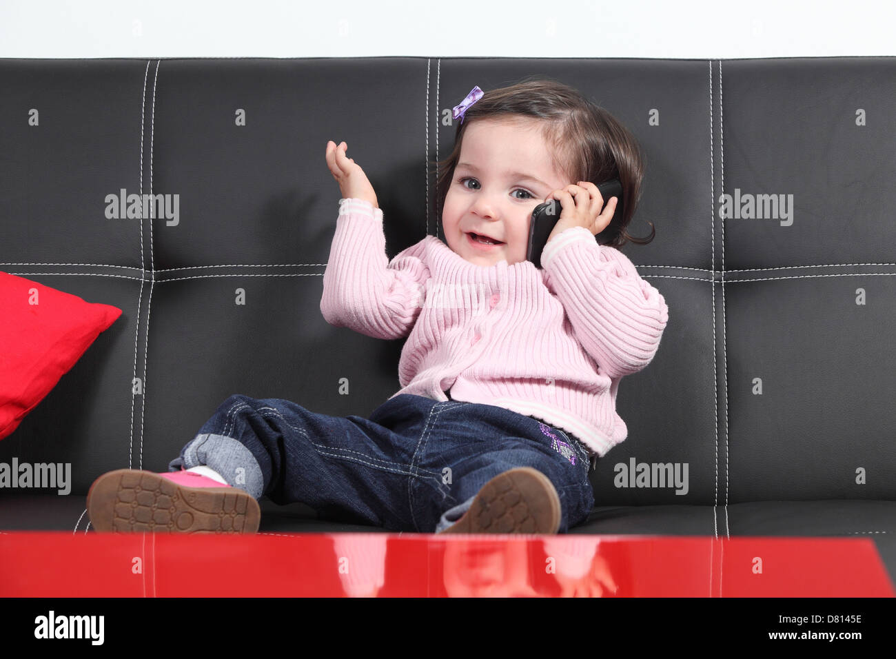 Casual baby giocando felice con un telefono cellulare seduta su un divano nero a casa Foto Stock