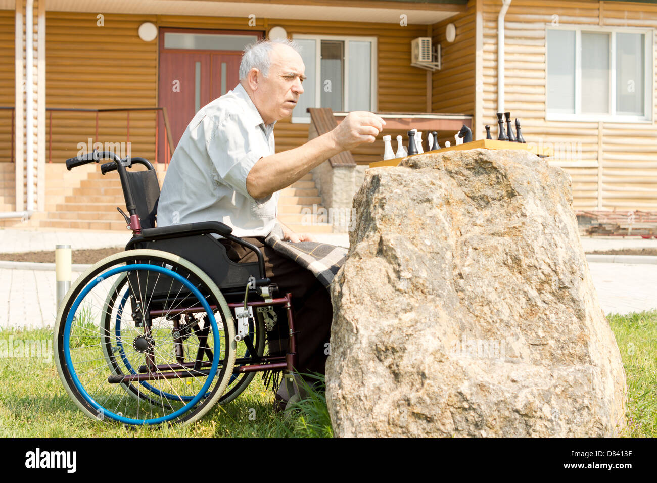 Disabilitato l'uomo gioca a scacchi nel suo giardino seduto nella sua sedia a rotelle con la scheda bilanciato su una roccia Foto Stock