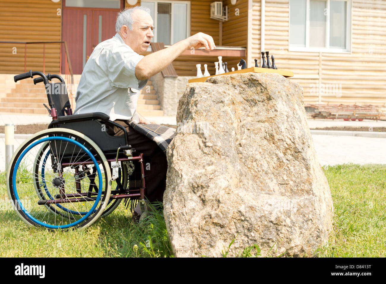 Anziani disabili uomo giocando a scacchi seduta nella sua sedia a rotelle davanti a casa sua nel giardino con la scacchiera sulla parte superiore di un conveniente rock. Foto Stock