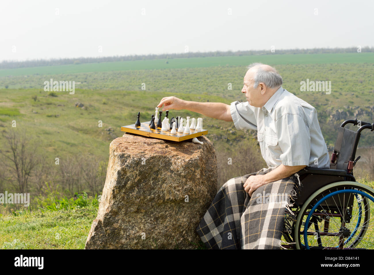 Uomo anziano in una sedia a rotelle a giocare a scacchi all'aperto con la scacchiera bilanciato su una roccia in una pittoresca campagna Foto Stock