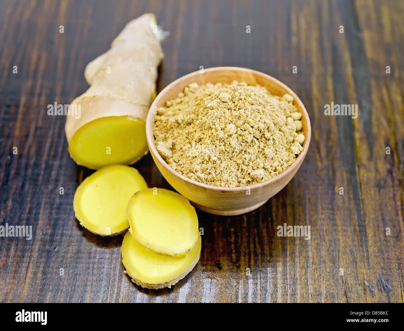 Lo zenzero in polvere in un recipiente di legno e fette di radice di zenzero al buio su un pannello di legno Foto Stock