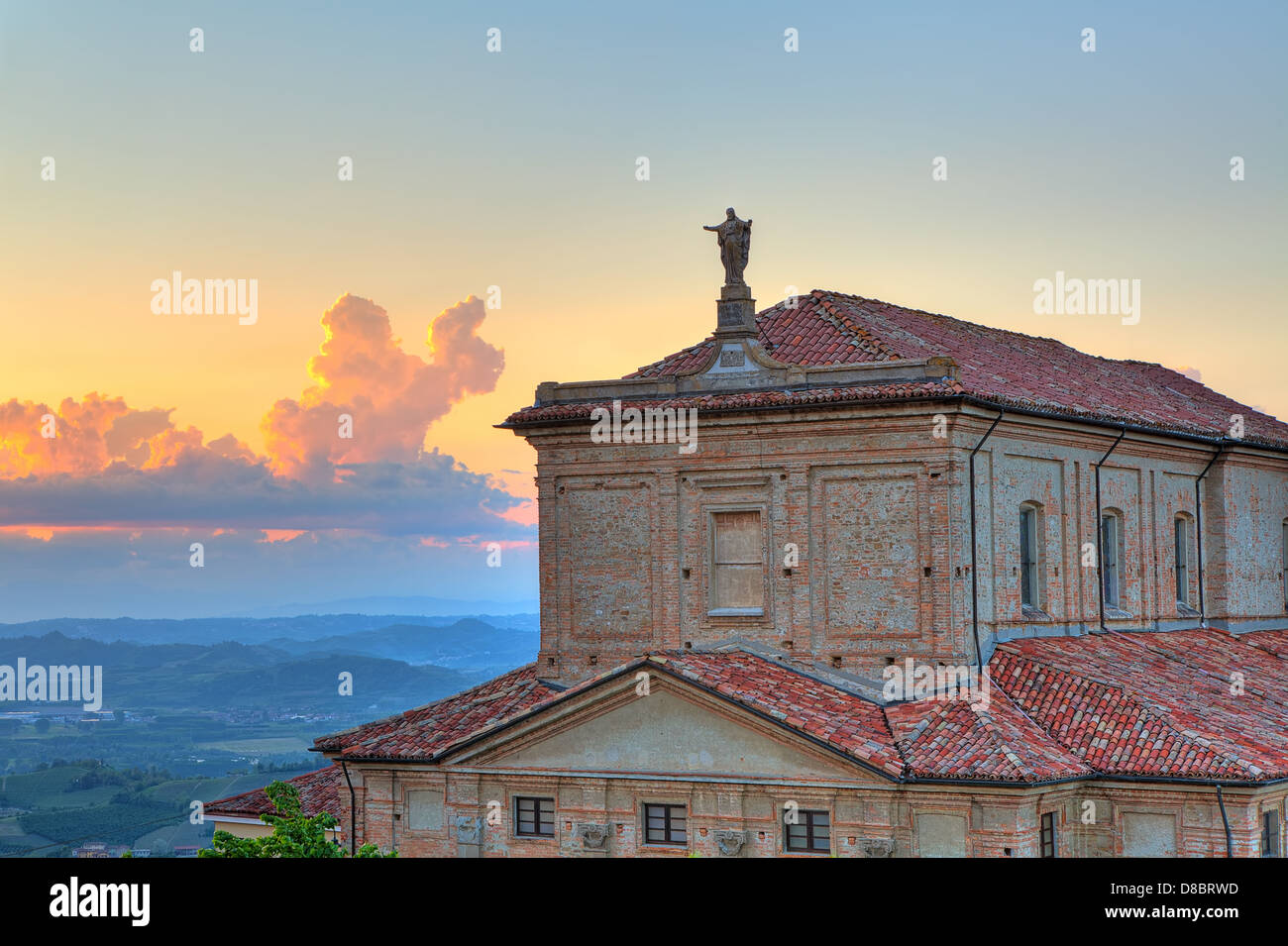 Statua di Gesù Cristo sul tetto della chiesa e le colline delle Langhe su sfondo al tramonto sotto il bellissimo cielo di sera in Italia. Foto Stock