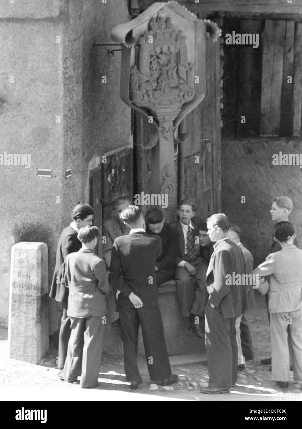 Un gruppo di adolescenti di sesso maschile - indossa una tuta come è comune negli anni sessanta, incontro presso la fontana nella piazza del paese durante il loro tempo libero. Foto Stock