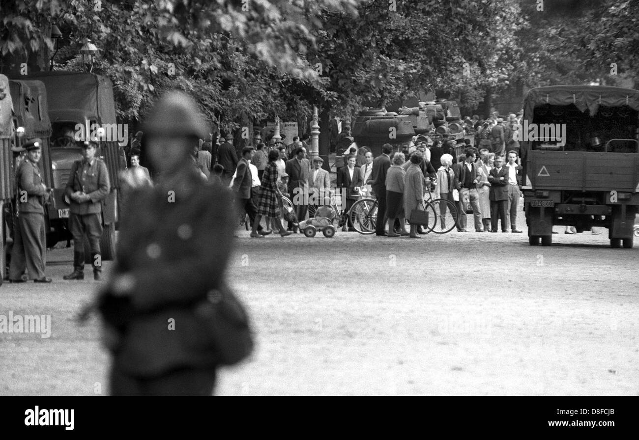 Un poliziotto GDR ("Volkspolizist') si erge di fronte ansiosi a Berlino Est i cittadini con la sua pistola sollevata su 'Swinemuender' Street a Berlino il borough di nozze, Berlino, agosto 1961. Sotto la protezione della RDT forze armate costruzione squadre hanno cominciato a errect blocchi stradali fatta di filo spinato al fine di barr fuori la parte orientale della città dalla western nelle prime ore del mattino del 13 agosto 1961. Il muro è stato destinato ad arrestare il numero sempre crescente di Oriente i berlinesi in fuga a Berlino Ovest. Foto Stock