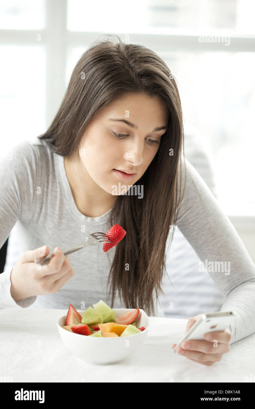 Ritratto di una giovane donna di mangiare insalata di frutta durante la navigazione con il suo iphone Foto Stock