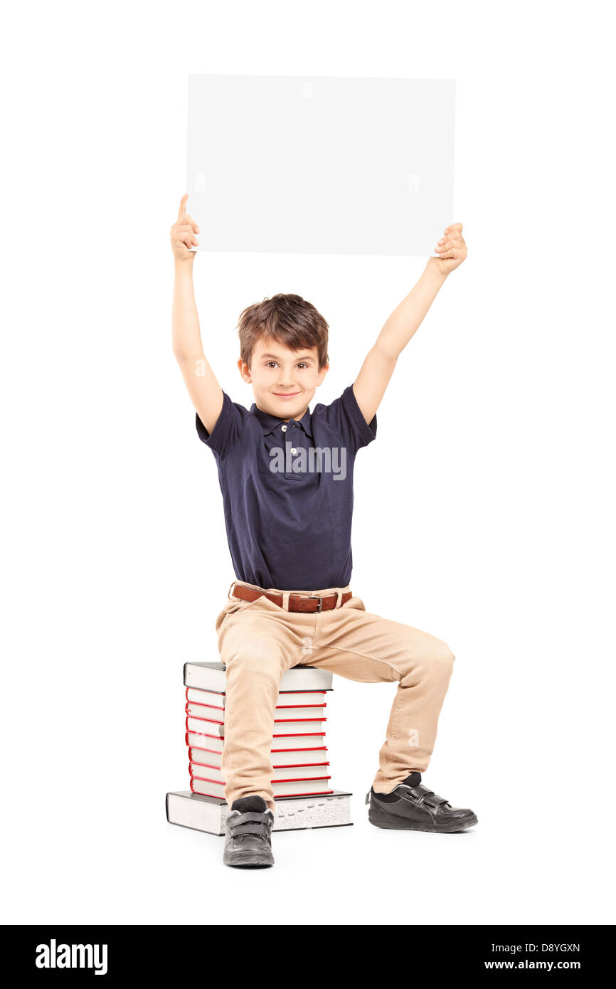 Felice ragazzo scuola tenendo un pannello vuoto sopra la sua testa, seduto su una pila di libri isolati su sfondo bianco Foto Stock