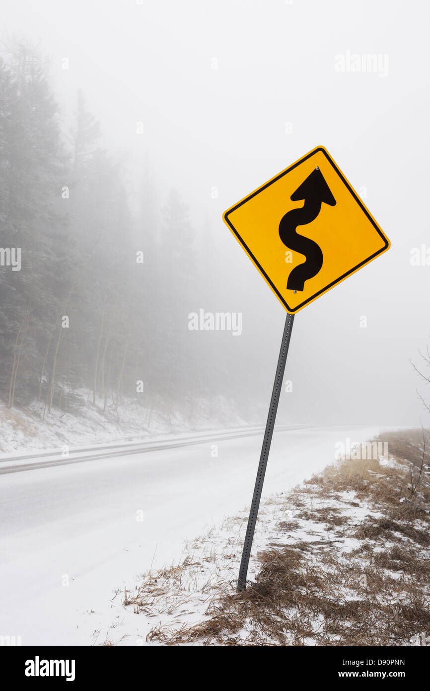 Segno di curva nella nebbia e neve su una strada di montagna. Foto Stock