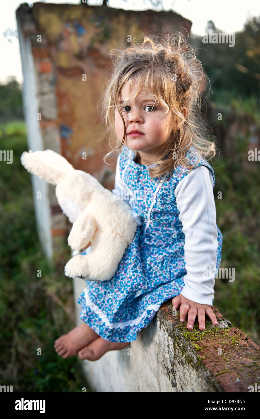 Giovani toddler con lunghi capelli biondi e fiorito di vestire seduto su una parete del vecchio edificio fatiscente tenendo un orsacchiotto di peluche. Foto Stock