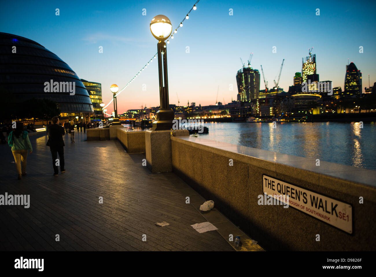 Queens Walk Municipio Skyline illuminato Crepuscolo Londra Regno Unito | LONDRA, Regno Unito — I pedoni passeggiano sul Queens Walk al crepuscolo oltre il Municipio di Londra sulla South Bank del Tamigi, con lo skyline illuminato della City di Londra che si staglia sull’acqua. Progettato da Norman Foster e inaugurato nel 2002, la forma sferica modificata dell’edificio minimizza la superficie per ridurre la perdita di calore. In costruzione a destra si trova il 20 Fenchurch Street, completato nel 2014. Foto Stock