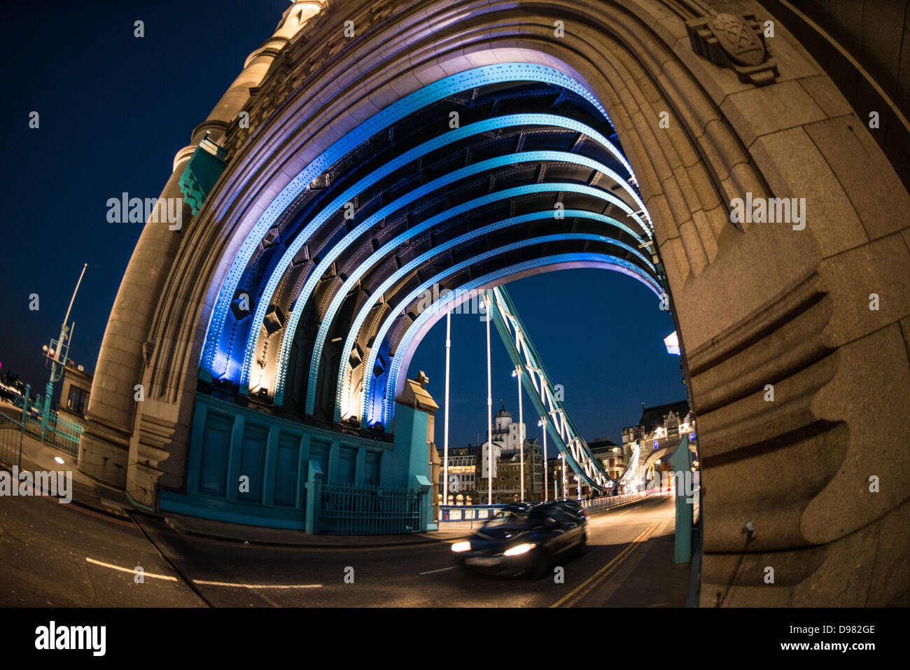 Arco illuminato del Tower Bridge Londra Regno Unito | LONDRA, Regno Unito — Costole d’acciaio illuminate di blu si incurvano sopra l’interno di una delle aperture ad arco di una torre del Tower Bridge, incorniciando un’auto di passaggio e la seconda torre del ponte oltre. La ferriera rivettata risale alla costruzione originale del 1886-1894, quando Sir William Arrol & Co. di Glasgow eresse lo scheletro in acciaio successivamente rivestito di granito della Cornovaglia e pietra di Portland. Lo schema di illuminazione blu e bianco risale alla livrea del Giubileo d’Argento del ponte del 1977. Foto Stock