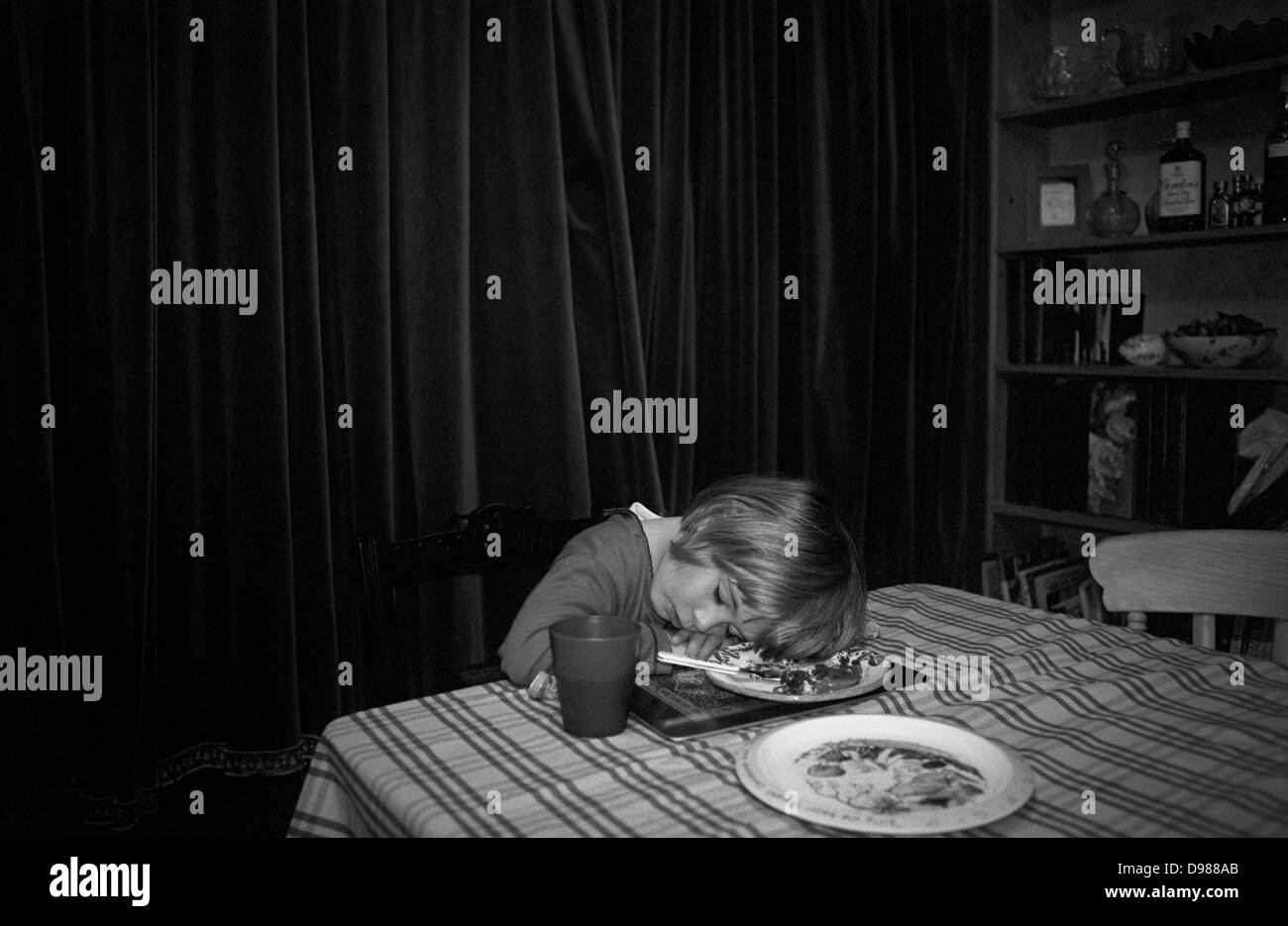 Un 3 anno-vecchia ragazza si addormenta prima di finire la sua cena e il cibo di cui è ancora sulla piastra sotto la sua testa, a casa nel suo sud casa di Londra. È presto la sera ed è stato un occupato, stancante giornata che ha terminato improvvisamente piuttosto al di sopra della piastra di cena. Pesanti tende di feltro sono disegnati per bloccare la notte d'inverno e i resti del suo tè e semi-bevuto bevande è sul tavolo panno. Foto Stock