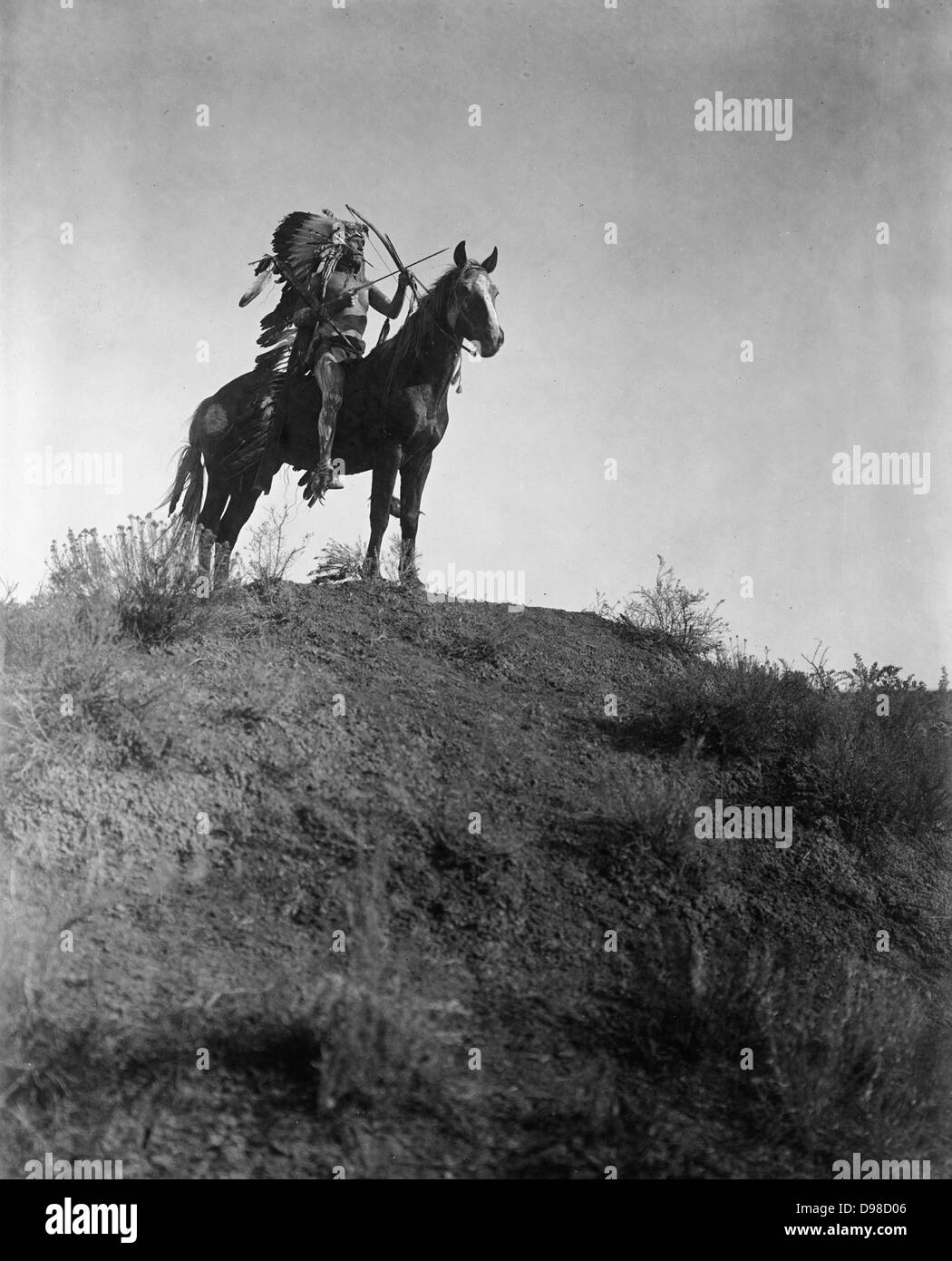 Native American Indian uomo in piuma copricapo, a cavallo, tenendo arco e frecce, 1 freccia nella sua bocca, 1908. Fotografia di Edward Curtis (1868-1952). Foto Stock