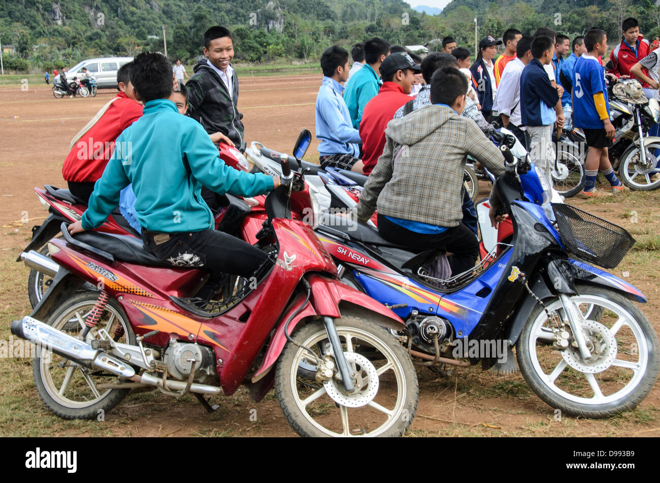 Studenti spettatori Scooters Crossbow Contest Vieng Xai Laos // VIENG XAI, Laos - gli spettatori degli studenti si allineano sui loro scooter e motociclette per assistere a una gara di balestra che si svolge su un campo di gioco sterrato nel terreno accidentato vicino a Vieng Xai nel nord-est del Laos. Il tradizionale evento sportivo attira partecipanti e spettatori dai villaggi circostanti in questa regione rurale montana. Vieng Xai, situata nella provincia di Houaphanh, è conosciuta per il suo paesaggio carsico calcareo e la rete di grotte che servirono come quartier generale per il Pathet Lao durante la seconda guerra d'Indocina. La città si trova vicino al bor Foto Stock
