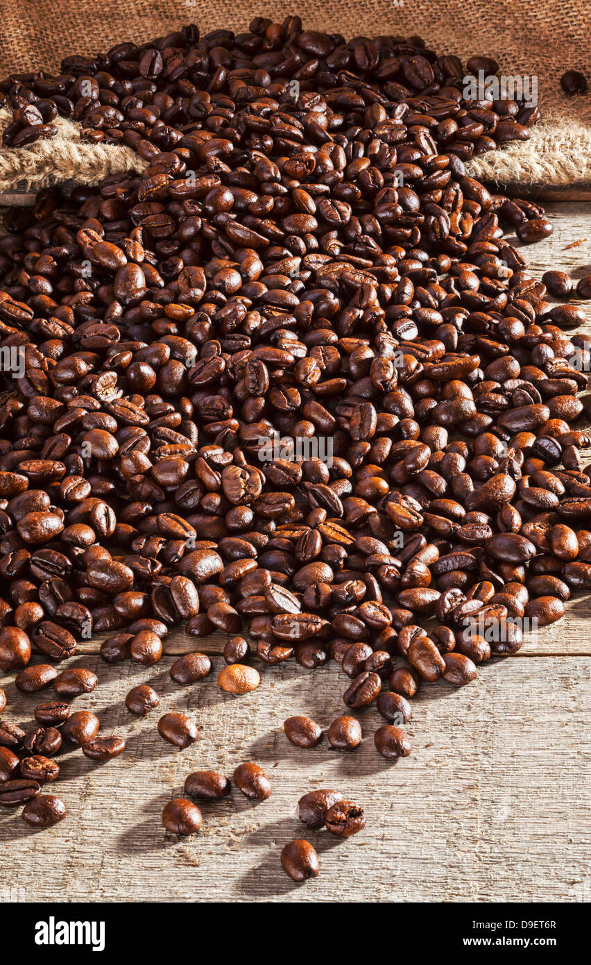 I chicchi di caffè su sfondo rustico - chicchi di caffè fuoriuscita da una tela o sacco di iuta su un listone rustico tavolo. Foto Stock