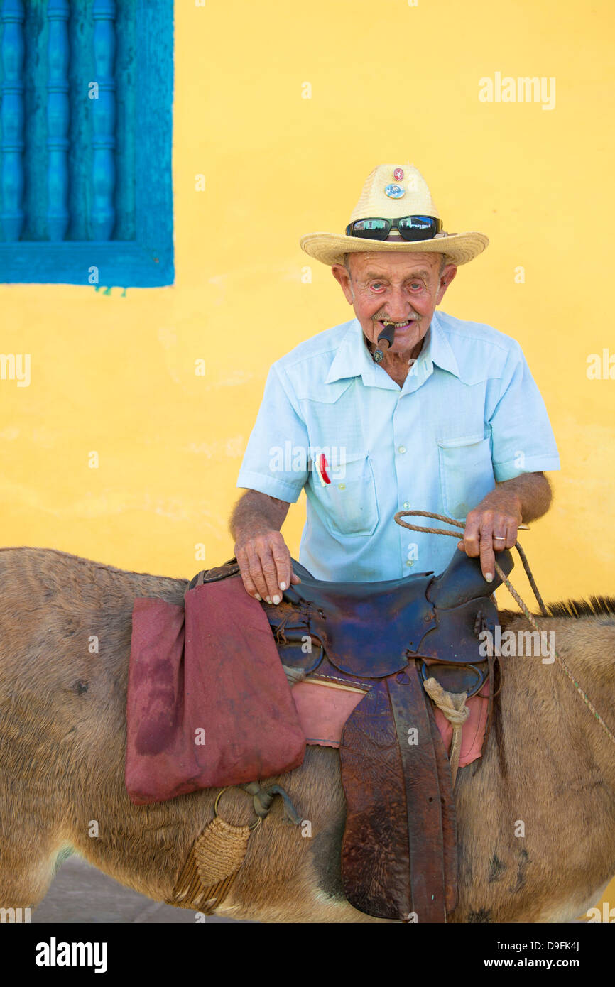 Ritratto di vecchio uomo di fumare un sigaro, posa contro una parete gialla con il suo asino per turisti di pesos, Trinidad, Cuba, West Indies Foto Stock