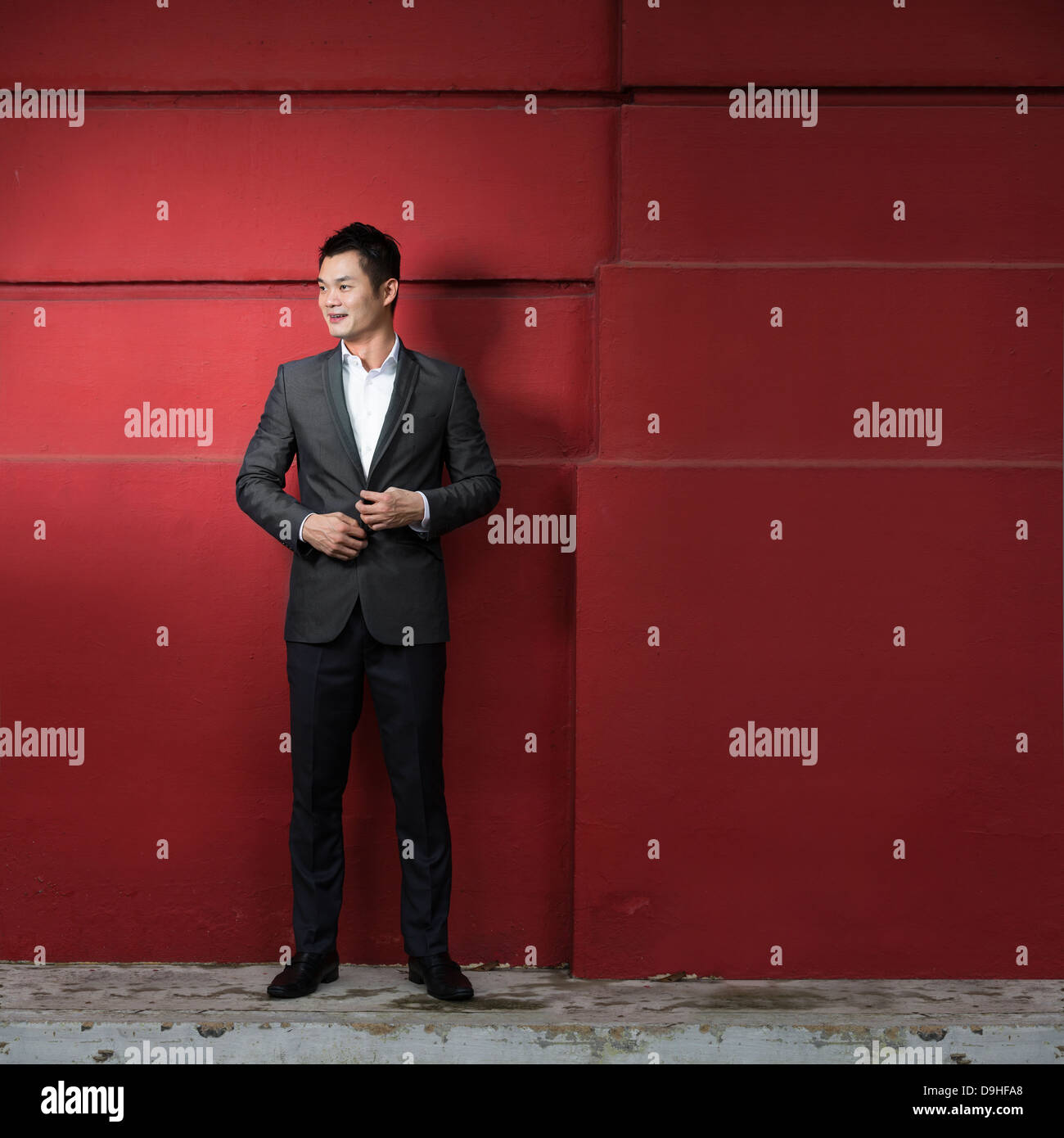 Bel giovane azienda cinese uomo appoggiato contro una parete rossa. Felice Asian business uomo sorridente e guardando lontano. Foto Stock