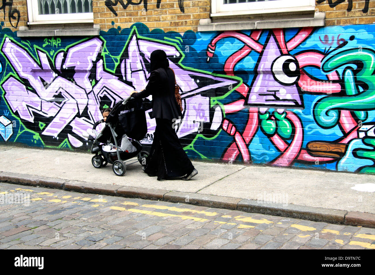 Donna musulmana con un contenuto della PRAM oltrepassando un coperto di graffiti parete in Shoreditch, East London Foto Stock