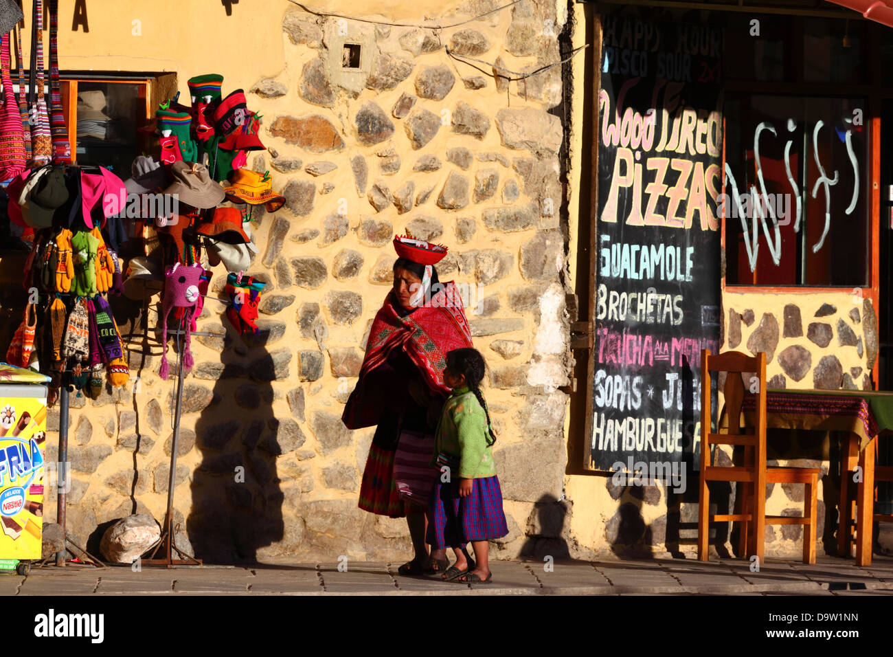 Signora indigena locale che indossa abiti tradizionali e ragazza che cammina accanto al negozio di souvenir e al menu del caffè, Ollantaytambo, Valle Sacra, vicino a Cusco, in Perù Foto Stock