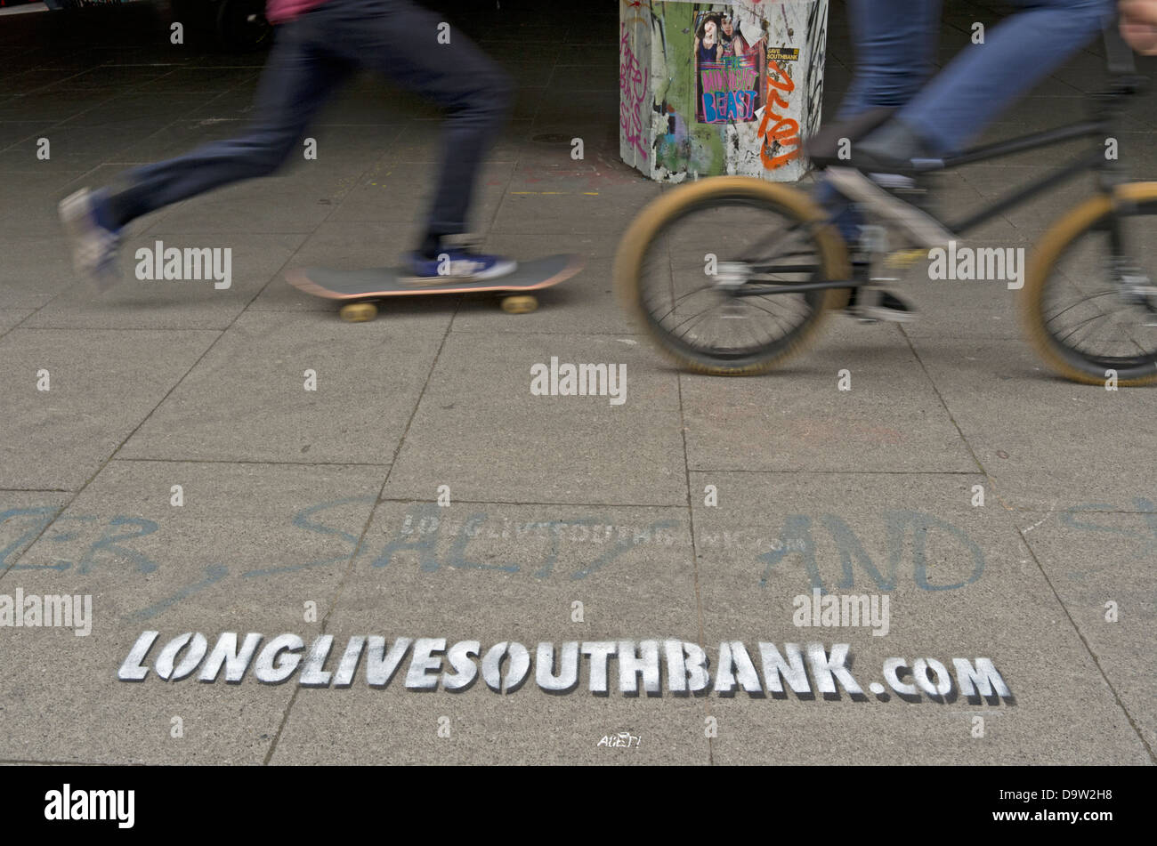 Un guidatore di skateboard e BMXer ride passato uno stencil promuovendo la lunga vita a South Bank skate park a Londra. Foto Stock